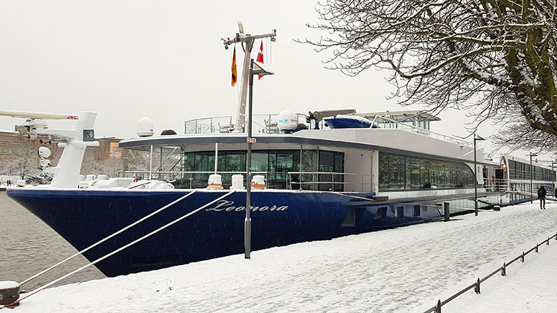 Elvecruisebåten MS Leonora ved kai på vinterstid. Promenade og trær med mye snø. 