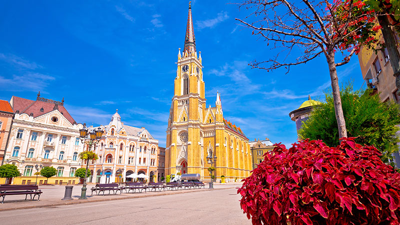 Stor kirke ved en pen plass i Novi Sad. Trr og benker i ulike farger. 