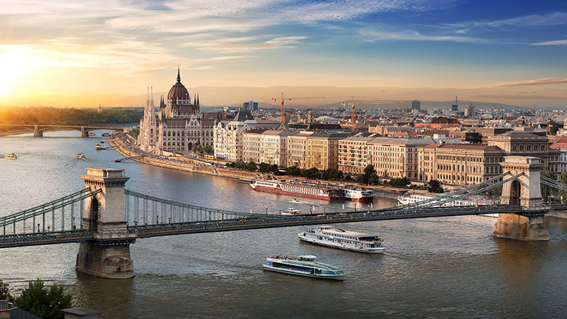 To elvecruise båter under stor bro i Budapest. Solnedgang over byen.