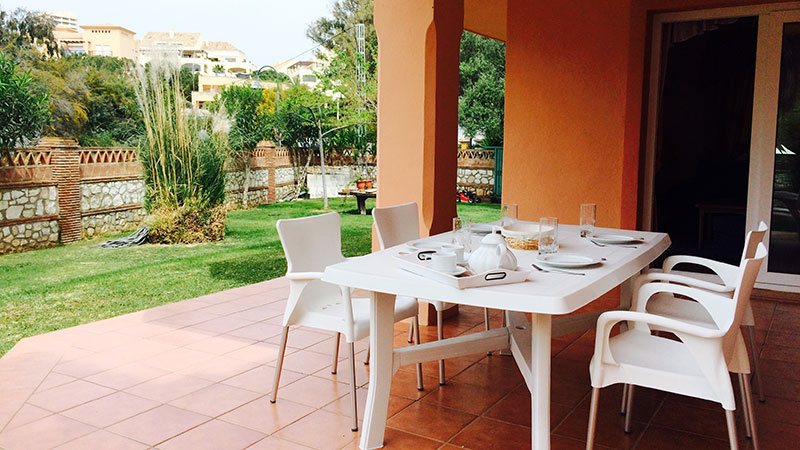 Spisebord og fire stoler på terasse utenfor leilighet. Grønn hage med palmer. 